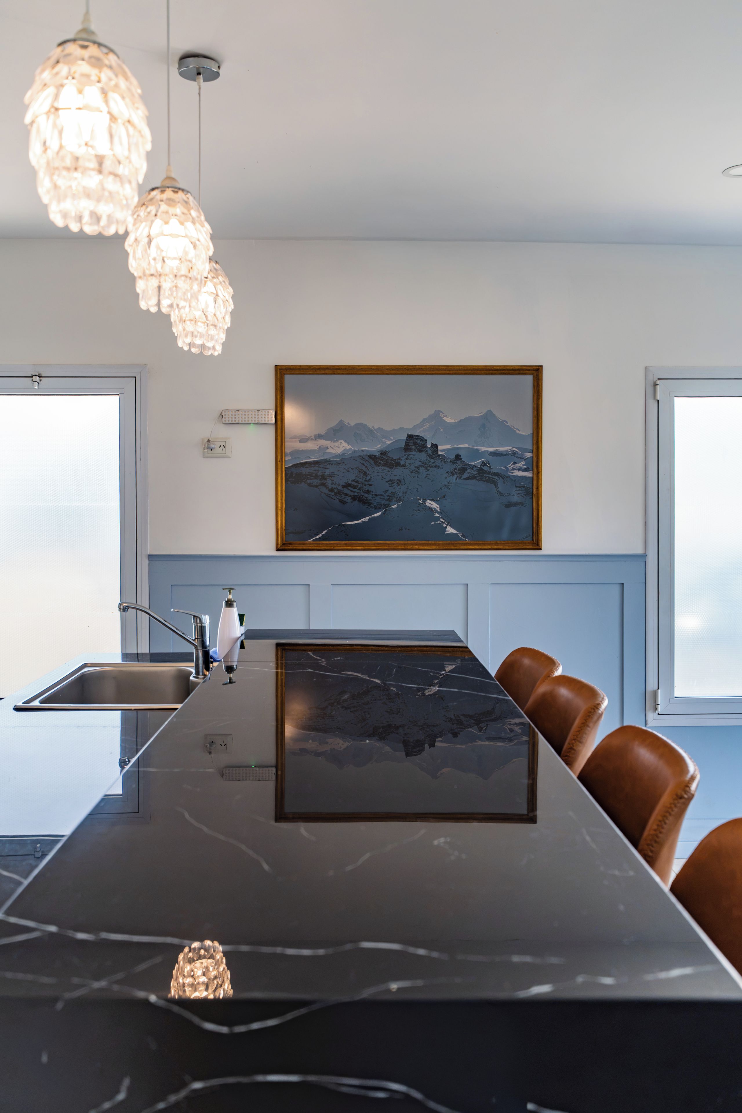 A modern kitchen with a marble island, pendant lights, a framed mountain landscape, and a window.