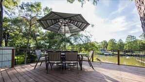 A wooden deck with a table and chairs under a large umbrella, overlooking a body of water and surrounded by trees.