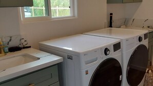 A laundry room with two front-loading washing machines, a sink, and a window with a view of greenery.