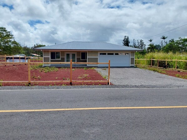 A single-story building with a garage, surrounded by a gravel driveway and a fenced area with plants. - Ainaloa Vacation Home of Hawaii! @3-bedroom house in tranquil Pāhoa with WiFi (Pahoa)