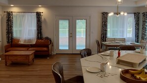 A spacious living area with a sofa, coffee table, and dining table. There is a ceiling fan, a window with curtains, and a door leading outside.