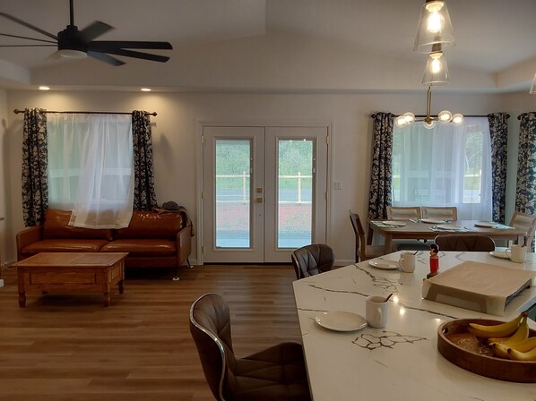 A spacious living area with a sofa, coffee table, and dining table. There is a ceiling fan, a window with curtains, and a door leading outside.