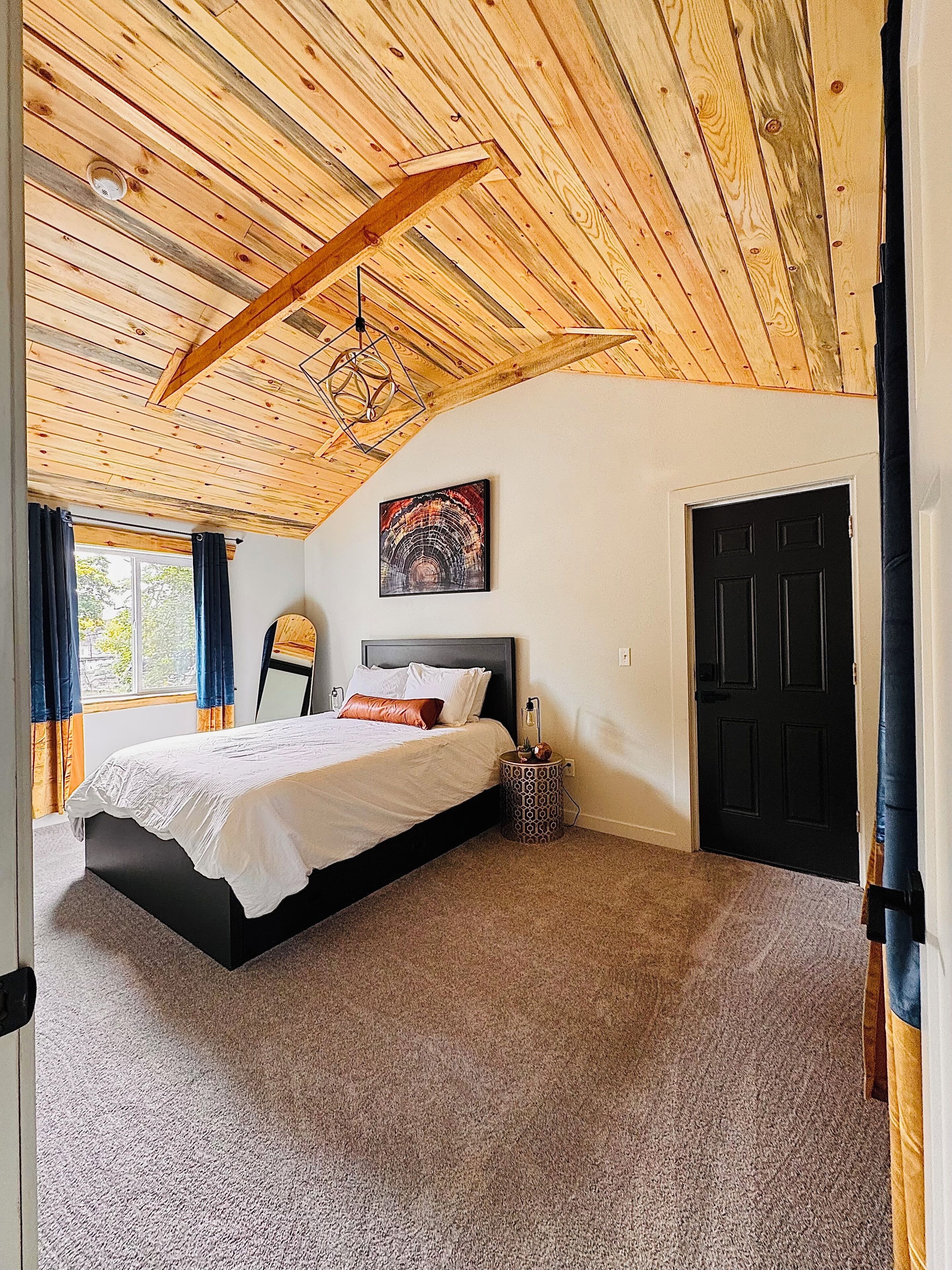 A bedroom with a wooden ceiling, a bed with white bedding, a nightstand with a lamp, and a black door.