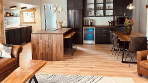A modern kitchen with dark cabinets, a central island, and bar stools. There is a dining area with a wooden table and chairs. - Luxury Living Near Downtown Moscow & U of I Campus (Moscow)