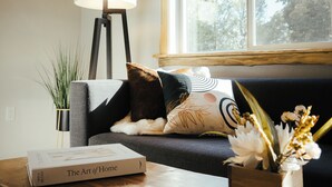 A cozy living room with a wooden ceiling, a sofa with decorative pillows, a floor lamp, a coffee table with a book titled "The Art of Home", and a window with a view of trees. - Luxury Living Near Downtown Moscow & U of I Campus (Moscow)