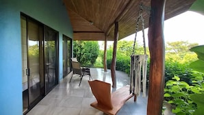 A covered outdoor area with a table and chairs, a hanging hammock, and a view of greenery.