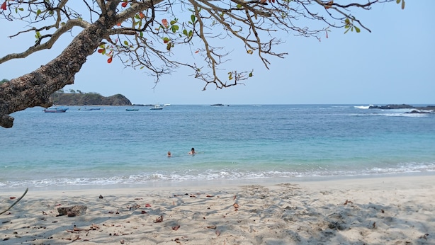 A beach with clear water, boats, and a rocky outcrop in the distance.
