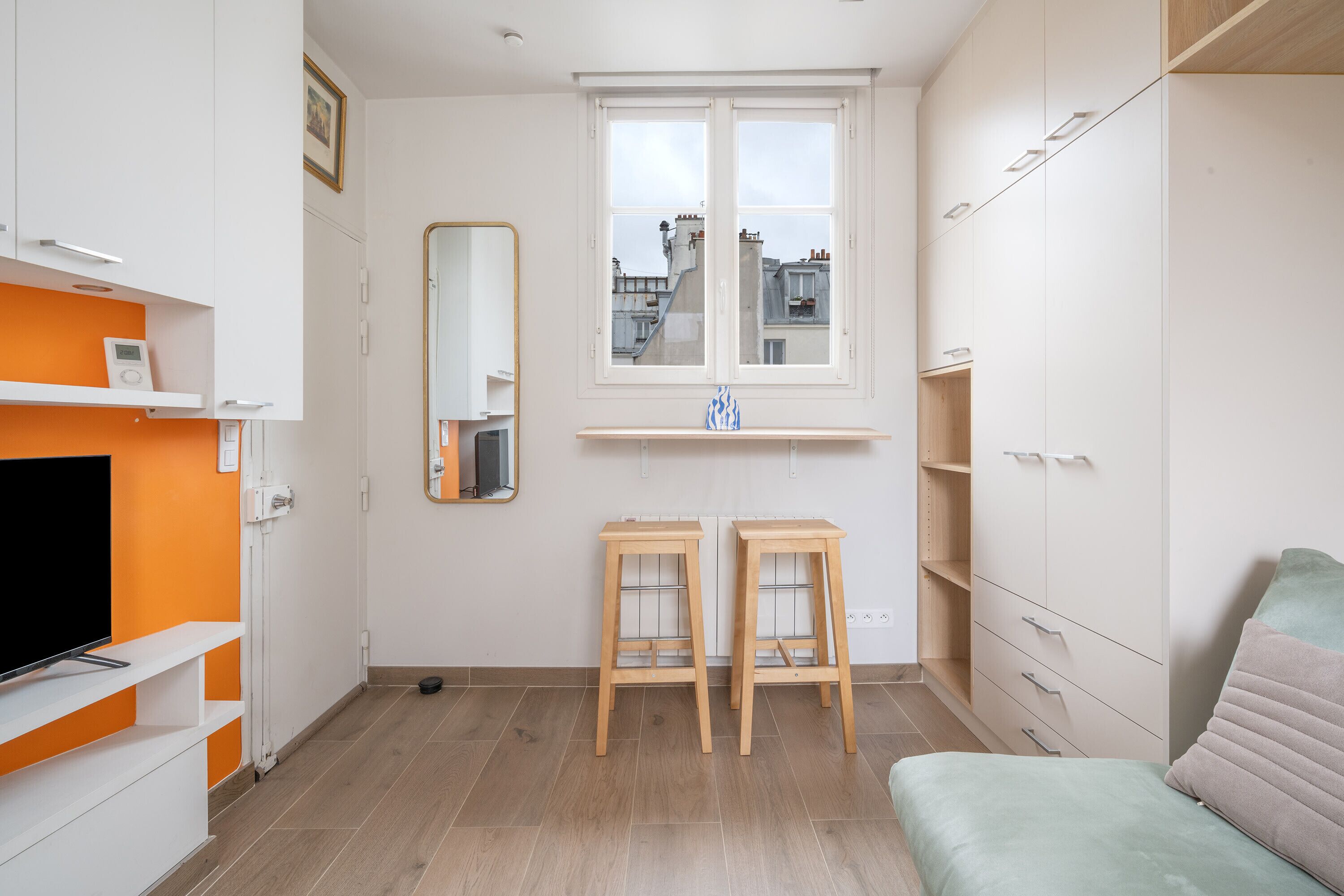 Une pièce moderne et bien éclairée, avec un téléviseur posé sur une étagère blanche, une fenêtre donnant sur des immeubles et un lit agrémenté d’un oreiller vert.