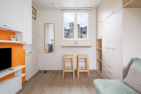 Una habitación moderna y bien iluminada con una televisión sobre una estantería blanca, una ventana con vistas a edificios y una cama con almohada verde.