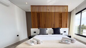 A modern hotel room with a large bed, wooden slats above the headboard, and a view of the city through large windows. - The Penthouse Gammarth (La Marsa)