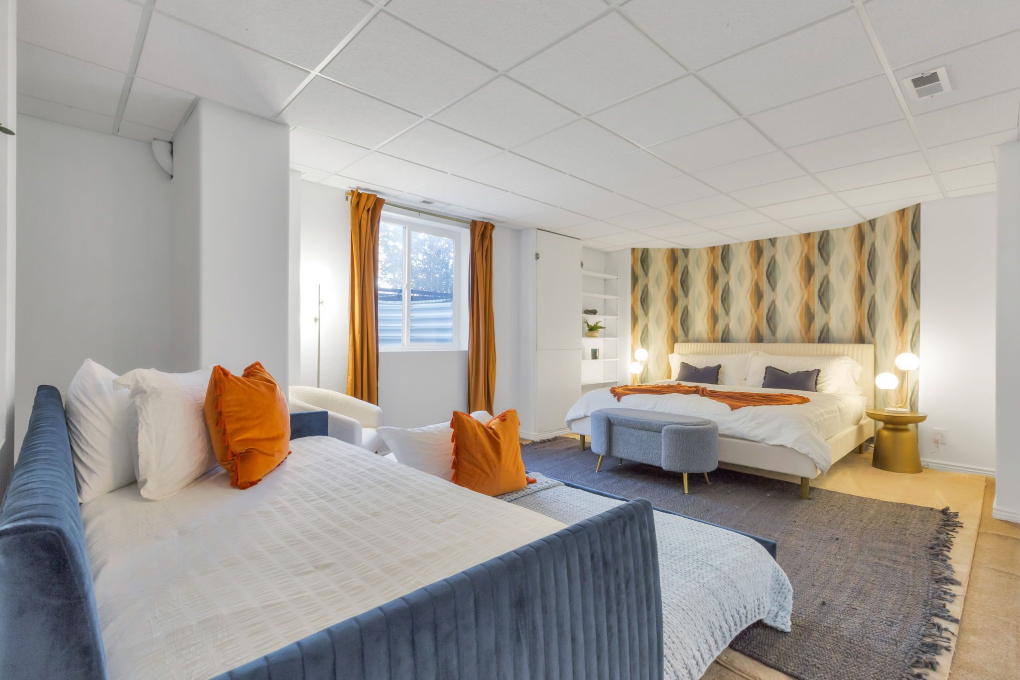 Moderni hotellihuone, jossa on suuri sänky, pienempi sänky, sohva ja työpöytä. Huoneessa on ikkuna, jossa on verhot, kuvioitu seinä ja yöpöytä, jossa on lamppu.