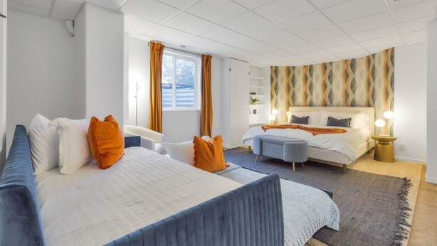 Habitación de hotel moderna con una cama grande, otra más pequeña, un sofá y un escritorio. Hay una ventana con cortinas, una pared con diseño decorativo y una mesita de noche con lámpara.
