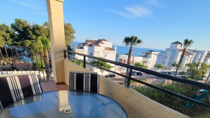 A round glass table with striped cushions, a balcony with a railing, and a view of buildings and trees.