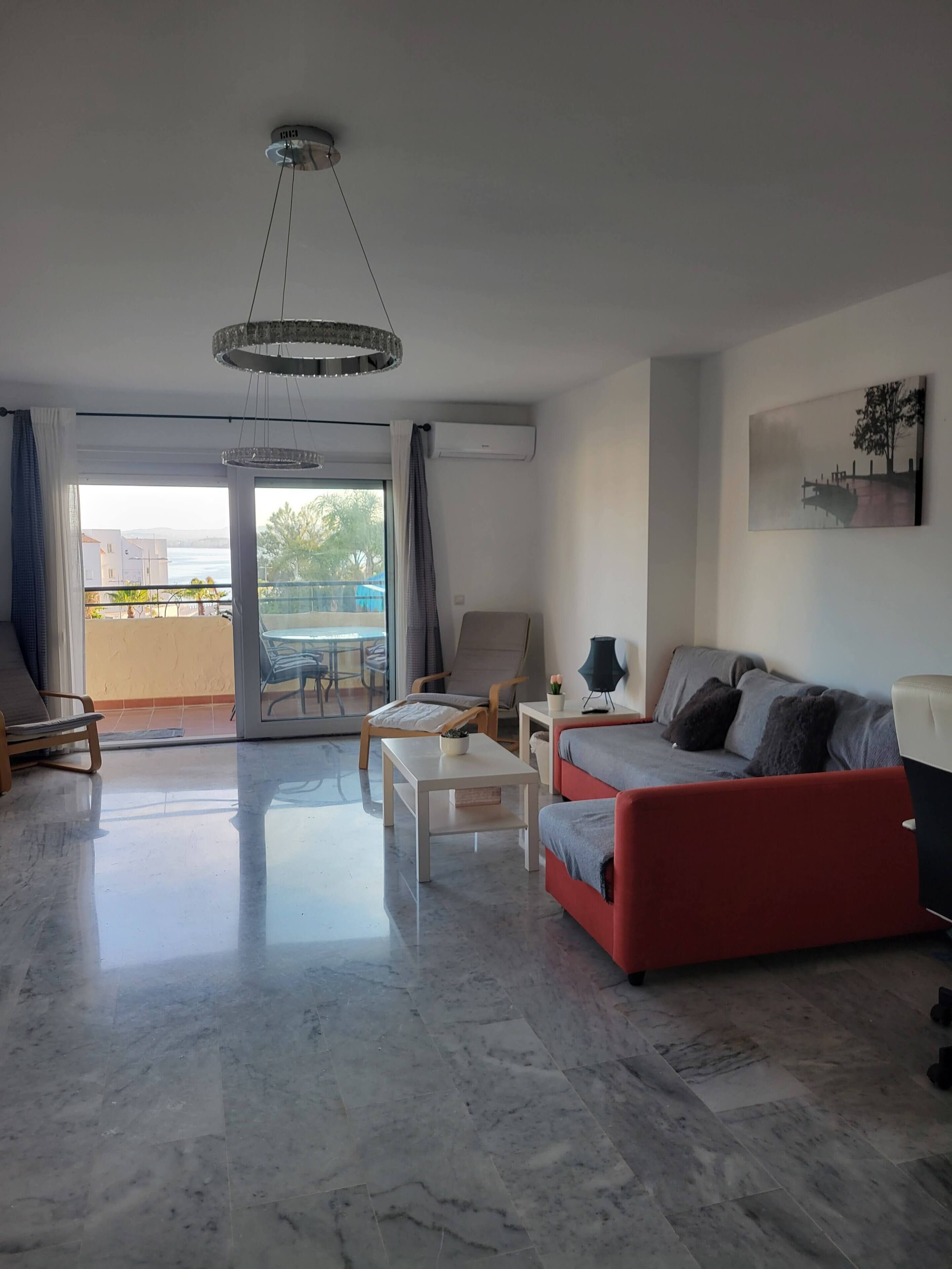 Ein modernes Wohnzimmer mit Sofa, Couchtisch und Sessel. Ein Balkon bietet Blick aufs Meer.