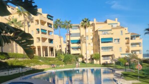 A swimming pool with a surrounding fence, palm trees, and a multi-story building in the background.