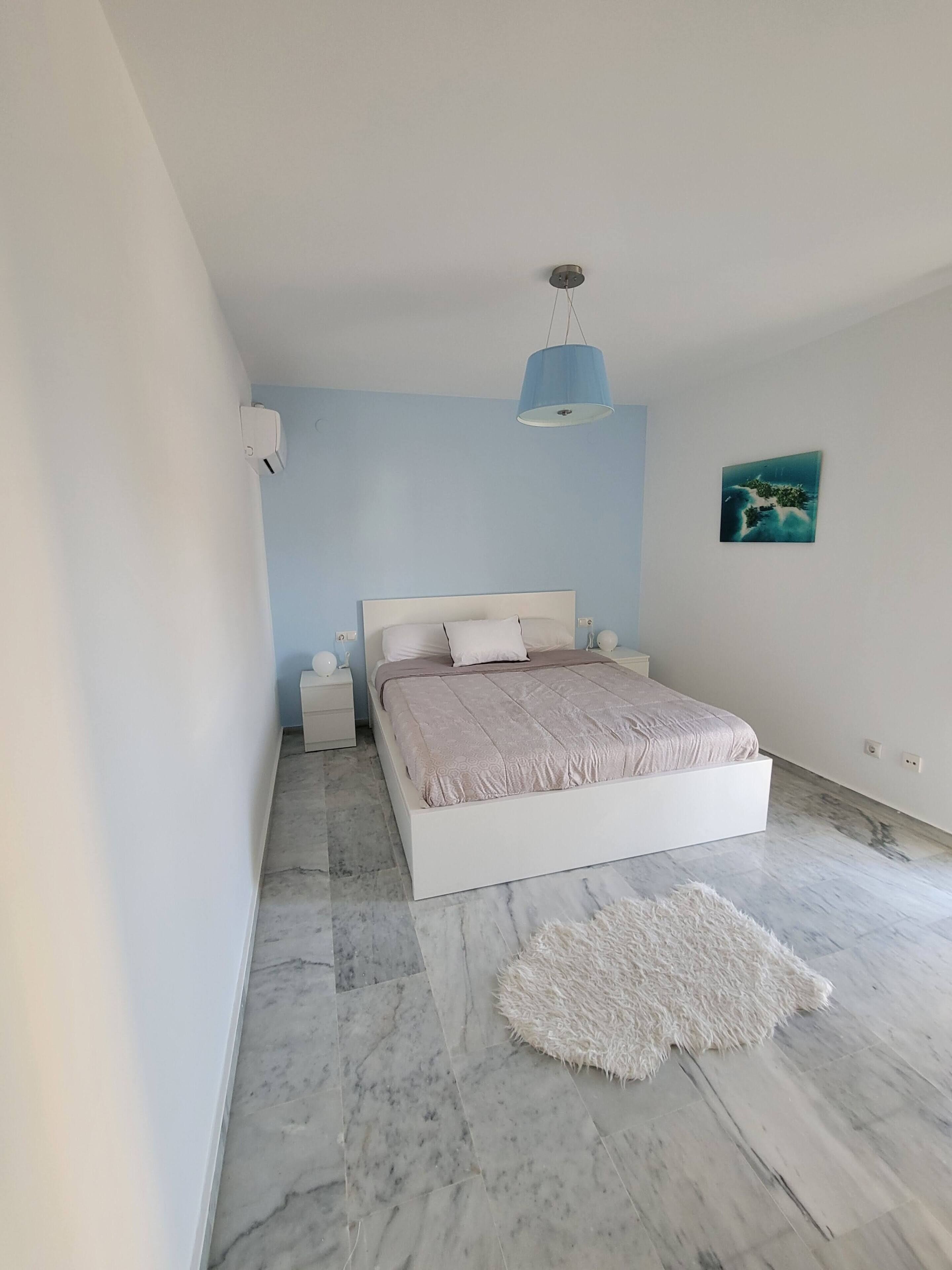 Un dormitorio con cama, mesitas de noche, alfombra blanca, lámpara colgante azul y una obra de arte en la pared.