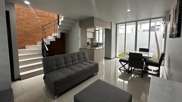 En moderne stue med en grå sofa, et spiseområde med bord og stole, et køkken samt en trappe til en etage over.