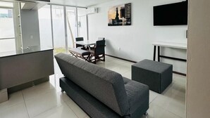 En moderne stue med en grå sofa, et spisebord med stole, et fladskærms-tv på væggen og et stort vindue, der lukker naturligt lys ind.