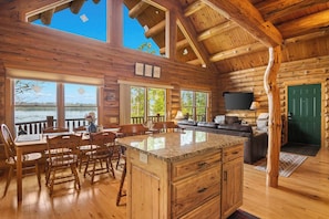 L’intérieur d’une cabane en rondins comprend un coin repas, une cuisine et un espace de vie, avec des murs en bois et de grandes fenêtres.