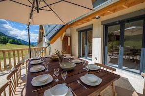 Un espace extérieur aménagé pour les repas, avec une table dressée pour six personnes, entourée de chaises en bois, d’un grand parasol et offrant une vue panoramique sur les montagnes.