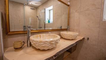A bathroom with two large, round, beige stone sinks on a wooden countertop, a gold-framed mirror, and a shower with a glass door.