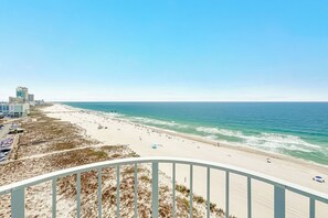 A beach view with a clear sky, a sandy beach, and a few buildings in the background.