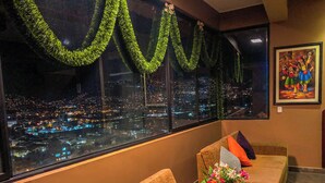 A hotel room with a large window overlooking a cityscape at night, furnished with two sofas and a small table with a vase of flowers.
