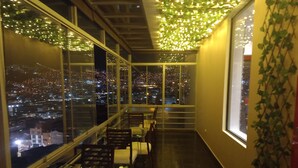A restaurant with a view of a cityscape at night, featuring tables and chairs arranged along a balcony.