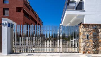 A gated entrance with a metal fence, a stone wall, and a modern building with balconies.