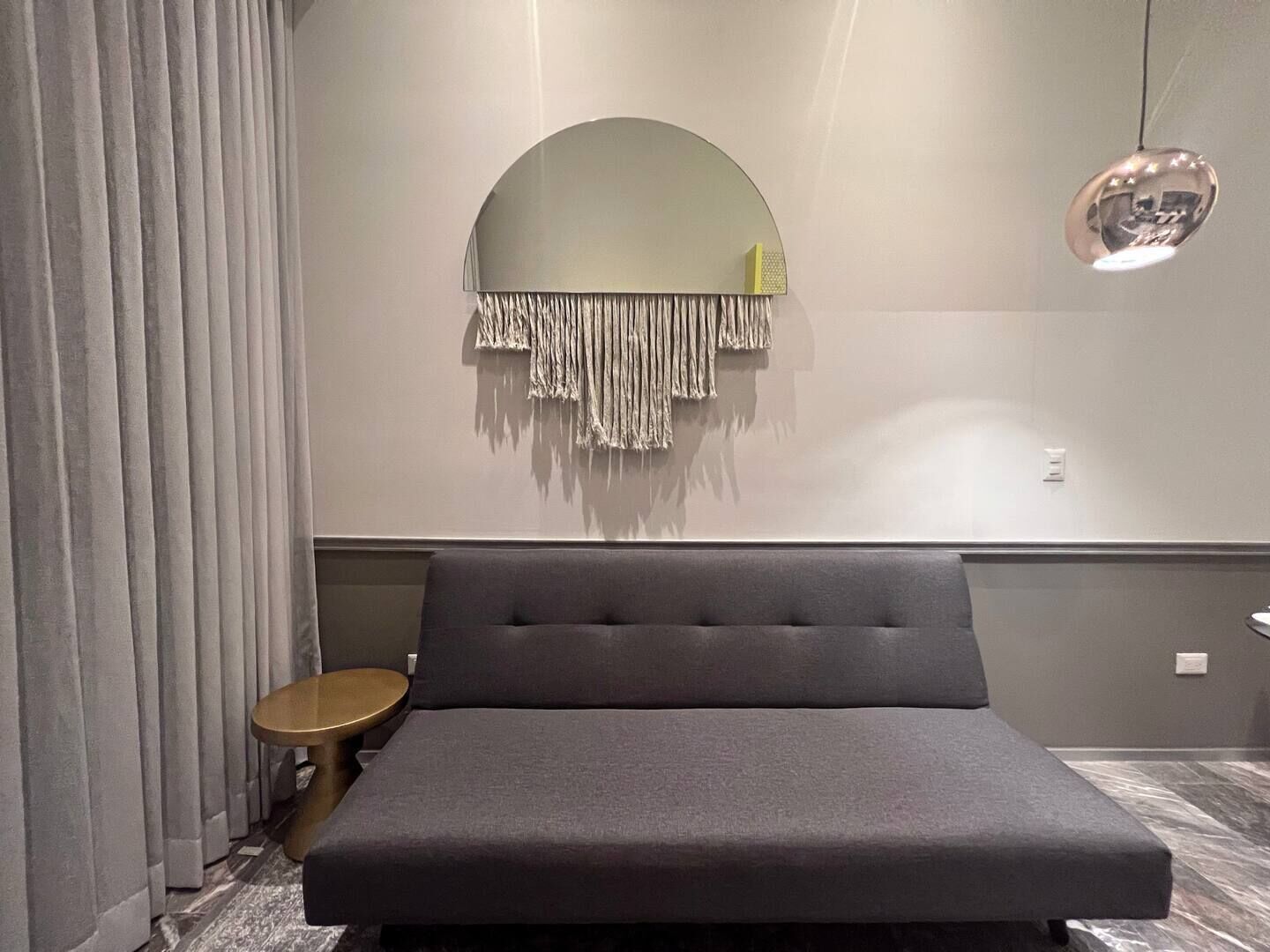 Un dormitorio moderno con un sofá cama gris, un espejo redondo con flecos y una pequeña mesa redonda.
