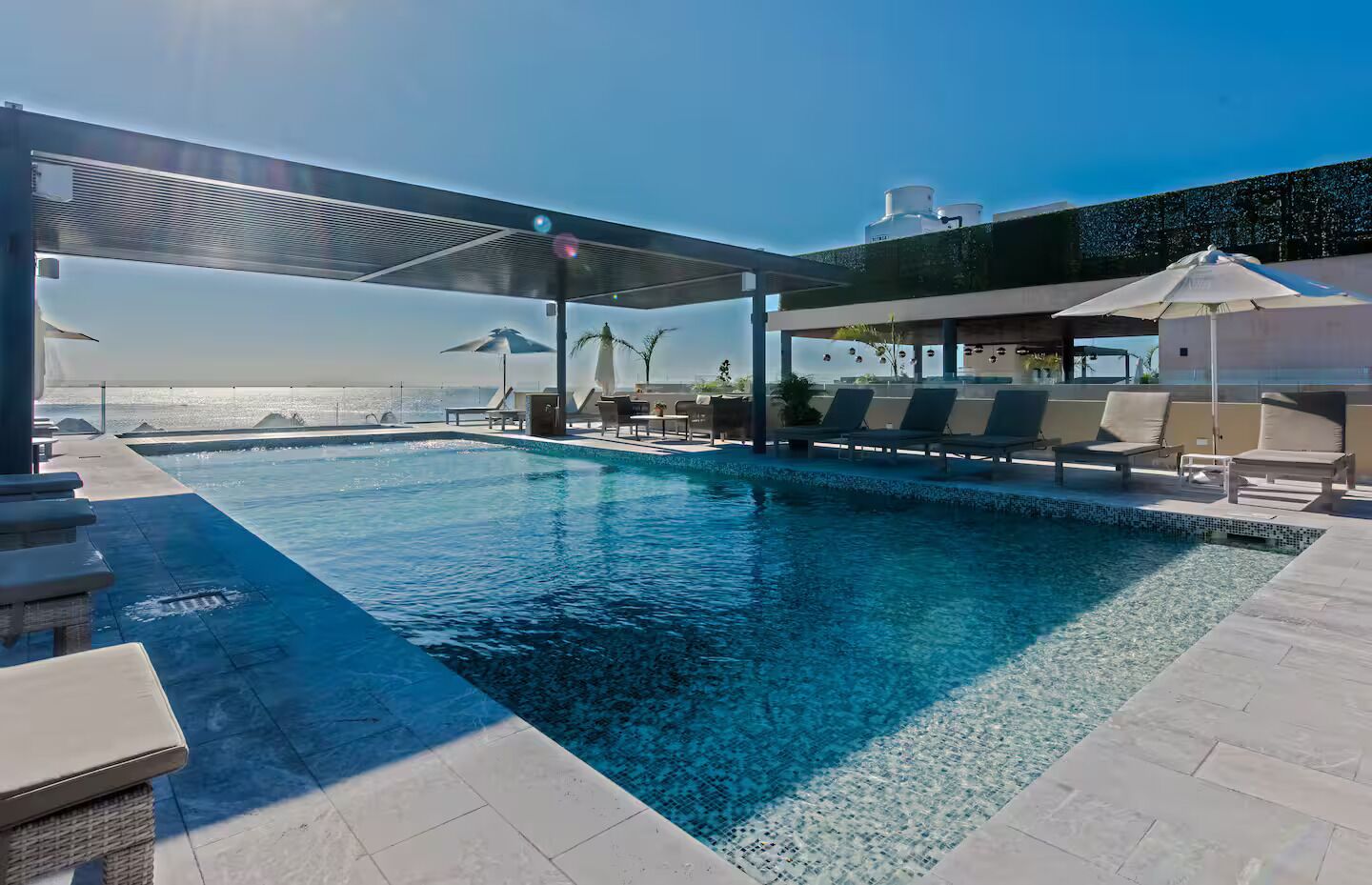 Piscina infinita en la azotea con sillas de descanso y sombrillas, con vista a una playa.