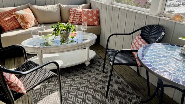 A sunlit outdoor seating area with a round table and chairs, a cushioned bench with patterned cushions, and a view of the sea through large windows.