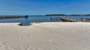 A beach with a wooden pier extending into calm water, a sandy area in the foreground, and a distant shoreline with greenery.