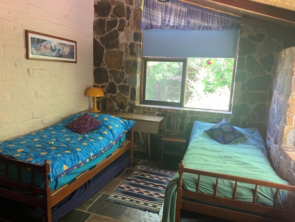 A room with two wooden beds, a desk, a chair, a window with a view of greenery, and a stone wall.