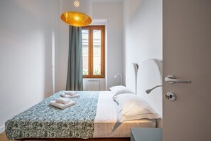 Un dormitorio con cama, mesita de noche, lámpara colgante, puerta y ventana.