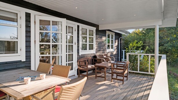 Une terrasse extérieure couverte, avec une table en bois et des chaises en osier, de grandes fenêtres et une vue sur la verdure.