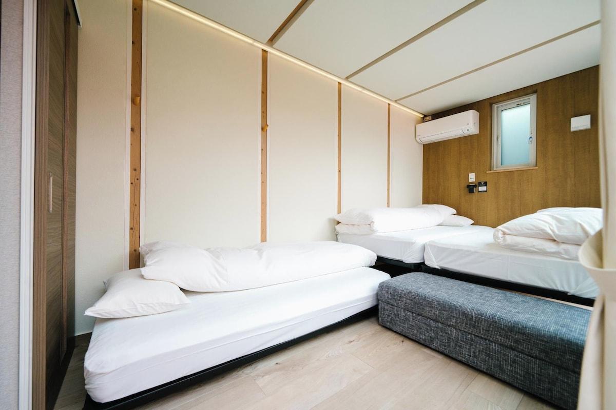 Una camera d'albergo compatta con due letti singoli, una piccola panca, un pannello di legno a parete e un condizionatore d'aria, oltre a una finestra.
