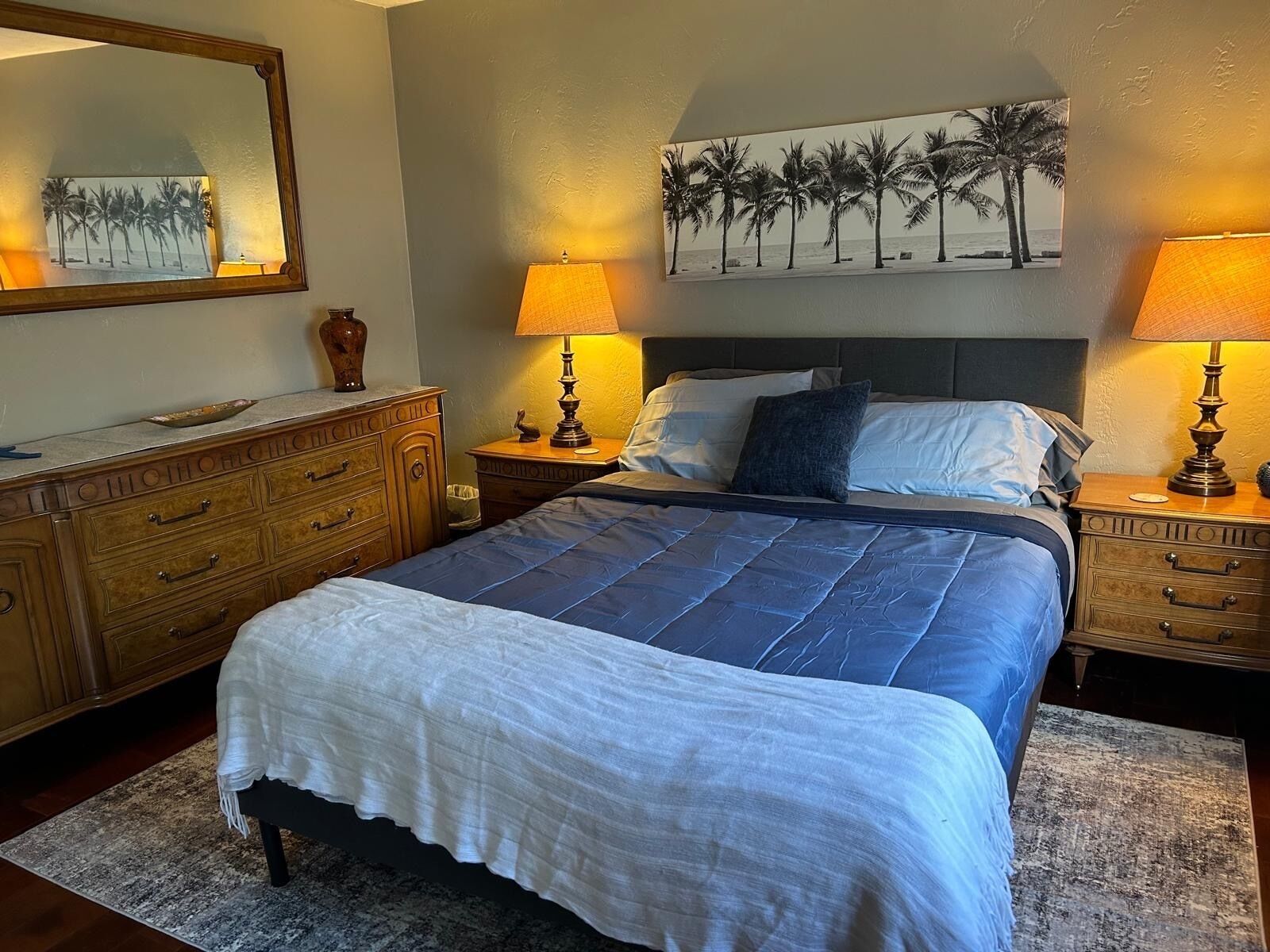 A bedroom with a large bed, two bedside tables with lamps, a dresser, a mirror, and a beach-themed painting on the wall.