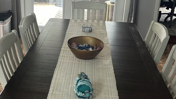A dining room with a dark wooden table, white chairs, a woven placemat, and a decorative bowl. - Blue Beach Pool House-Port Orange/Daytona (Port Orange)