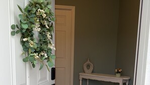 Un passadís amb una porta blanca, una corona decorativa, una taula de consola blanca amb un ram i una catifes amb disseny.