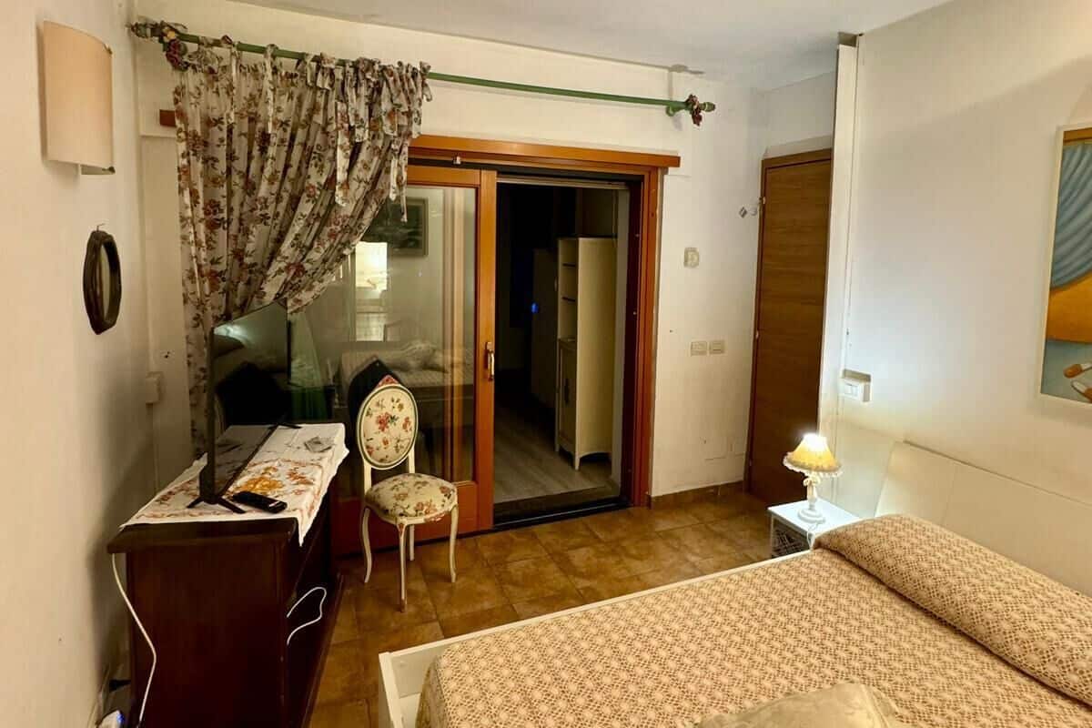 Quarto com cama, escrivaninha, cadeira, abajur e porta de vidro deslizante que dá para um corredor.