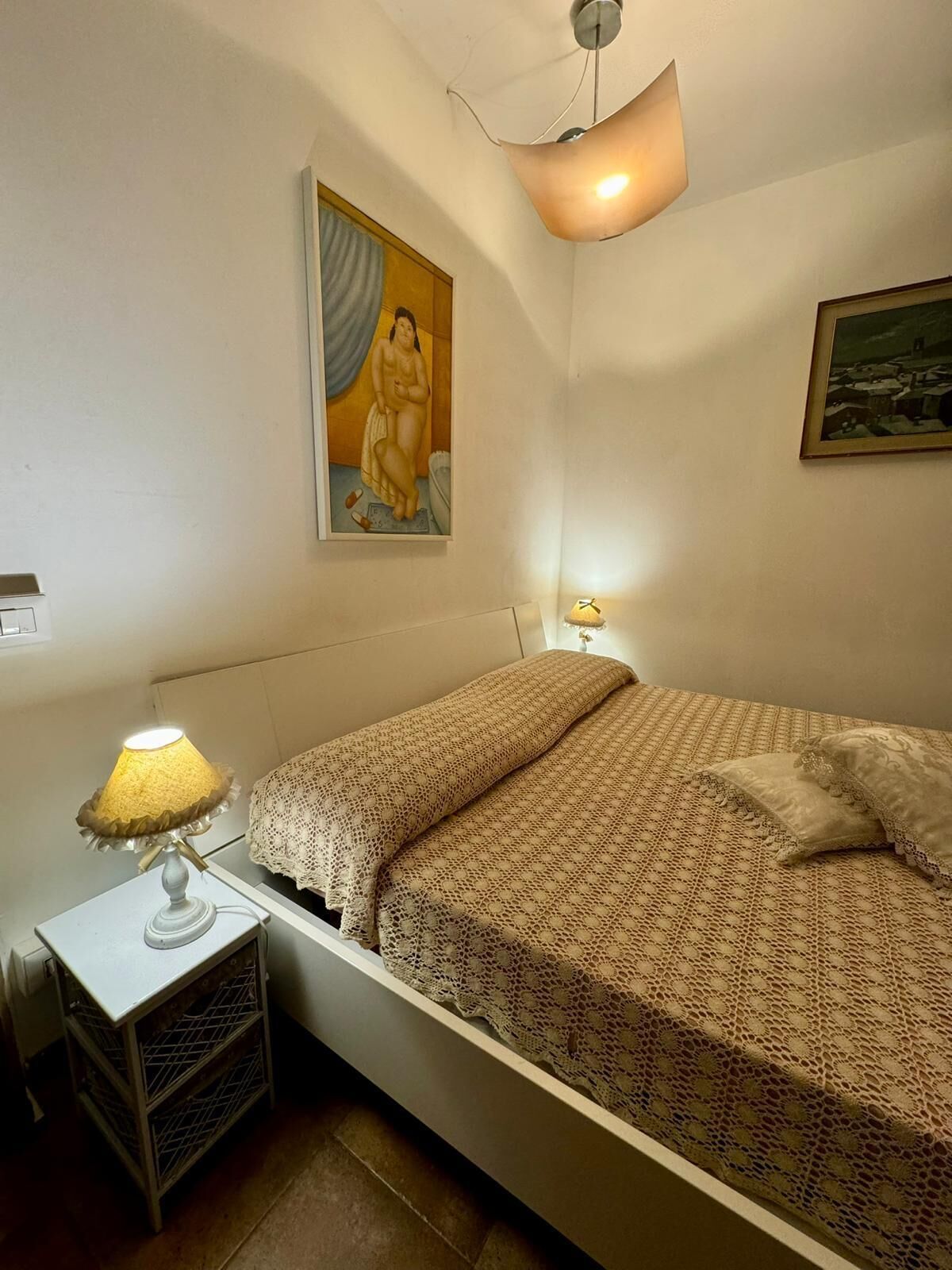 A bedroom with a bed, a nightstand with a lamp, a framed picture on the wall, and a ceiling light fixture.