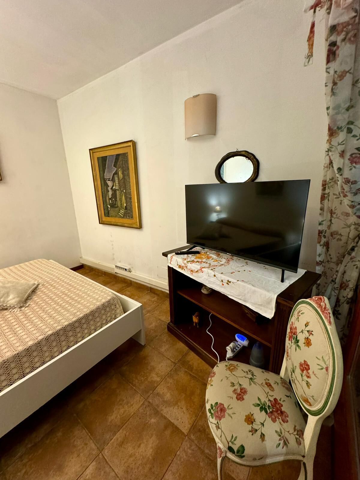 Een kleine, gemeubileerde kamer met een bed, een televisie op een standaard, een stoel en een wandlamp.