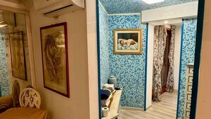 A room with a wooden floor, a yellow chair, a small table with a towel and a picture frame, and a wall with a blue mosaic pattern. -  L'affittacamere nel parco "Appia Antica" (Rome)