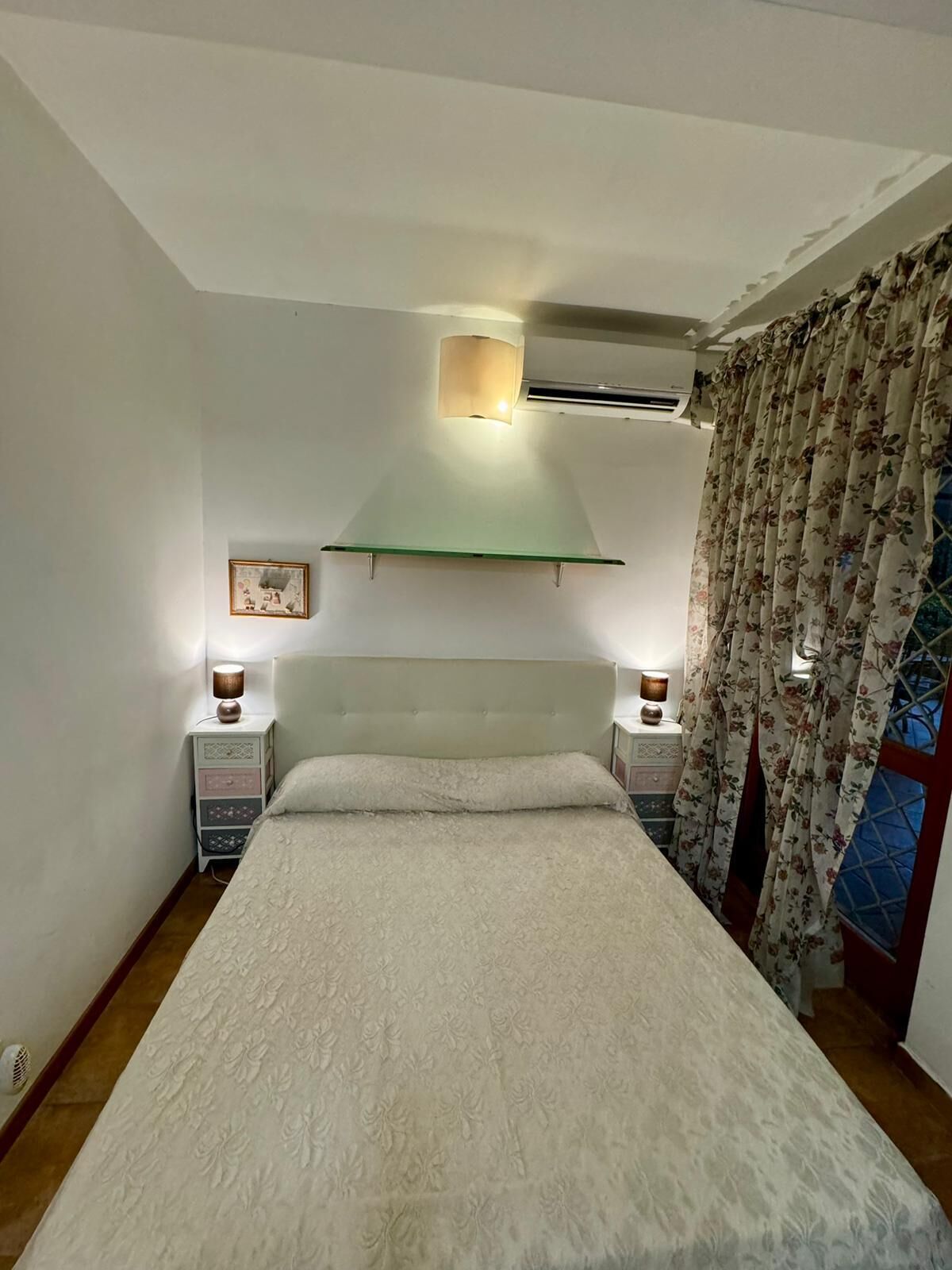 Ein Schlafzimmer mit einem Bett, zwei Nachttischen, einem Wandregal, einer Klimaanlage und einem Fenster mit floralen Vorhängen.