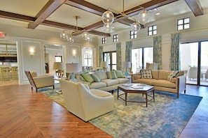 A spacious living room with a wooden floor, a beige sofa set, a green armchair, and a coffee table. There are multiple windows with curtains, a chandelier, and a visible exit sign. - Solara Resort home, private pool 6BR,4.5BA (Kissimmee)