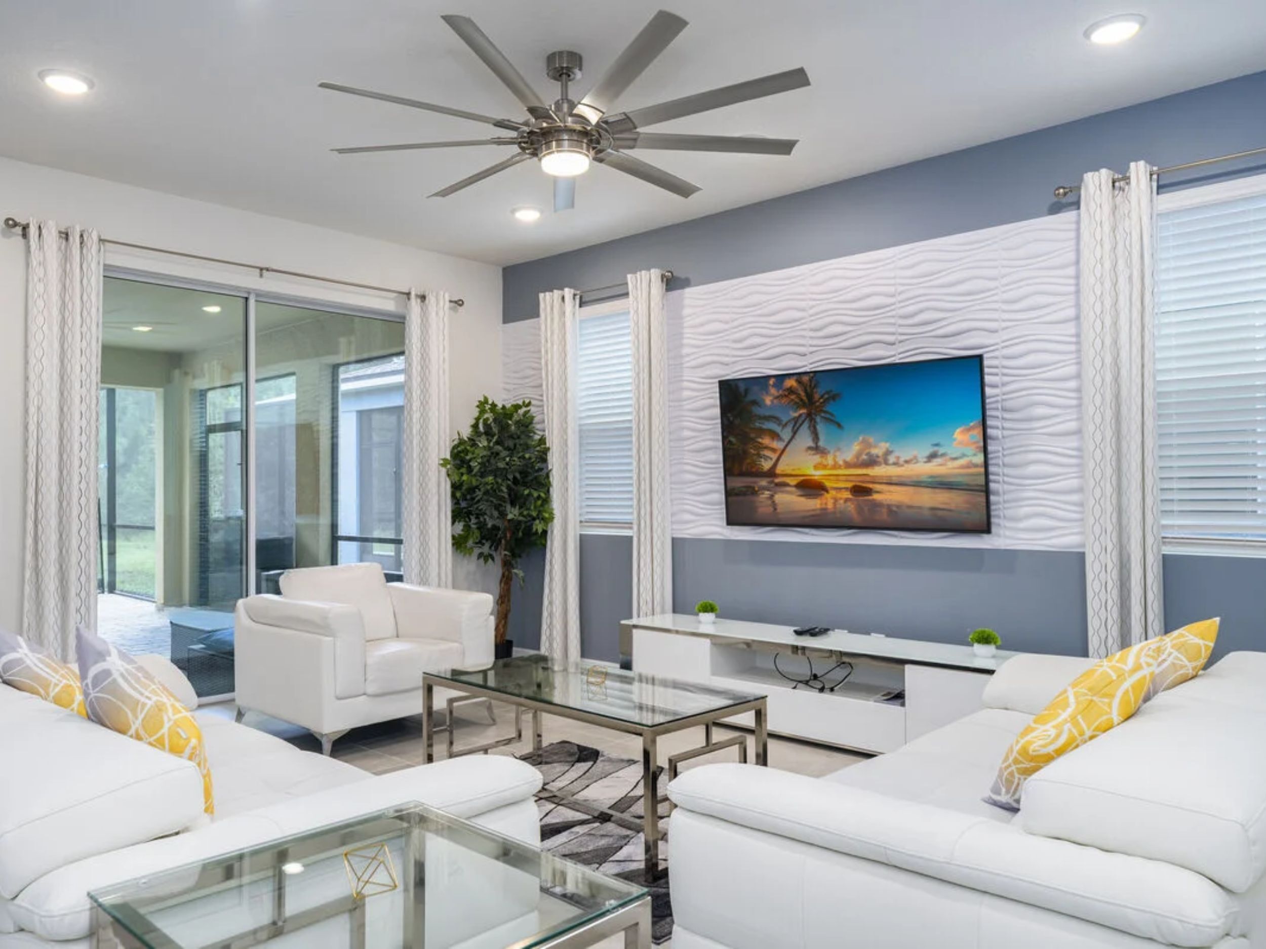 A modern living room with a ceiling fan, white sofas, a glass coffee table, and a large painting of a beach scene.
