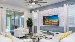 Ein modernes Wohnzimmer mit Deckenventilator, weißen Sofas, einem Glastisch und einem großen Gemälde mit Strandmotiv.