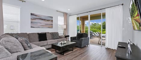 Una sala de estar moderna con sofá, sillones, una mesa de centro y televisor. Se aprecia un ventanal con cortinas y vistas al exterior con vegetación.