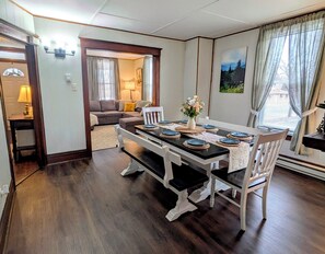Een eethoek met een houten tafel gedekt voor vier personen, witte stoelen en een vaas met bloemen. Achtergrond toont een bank en een lamp.
